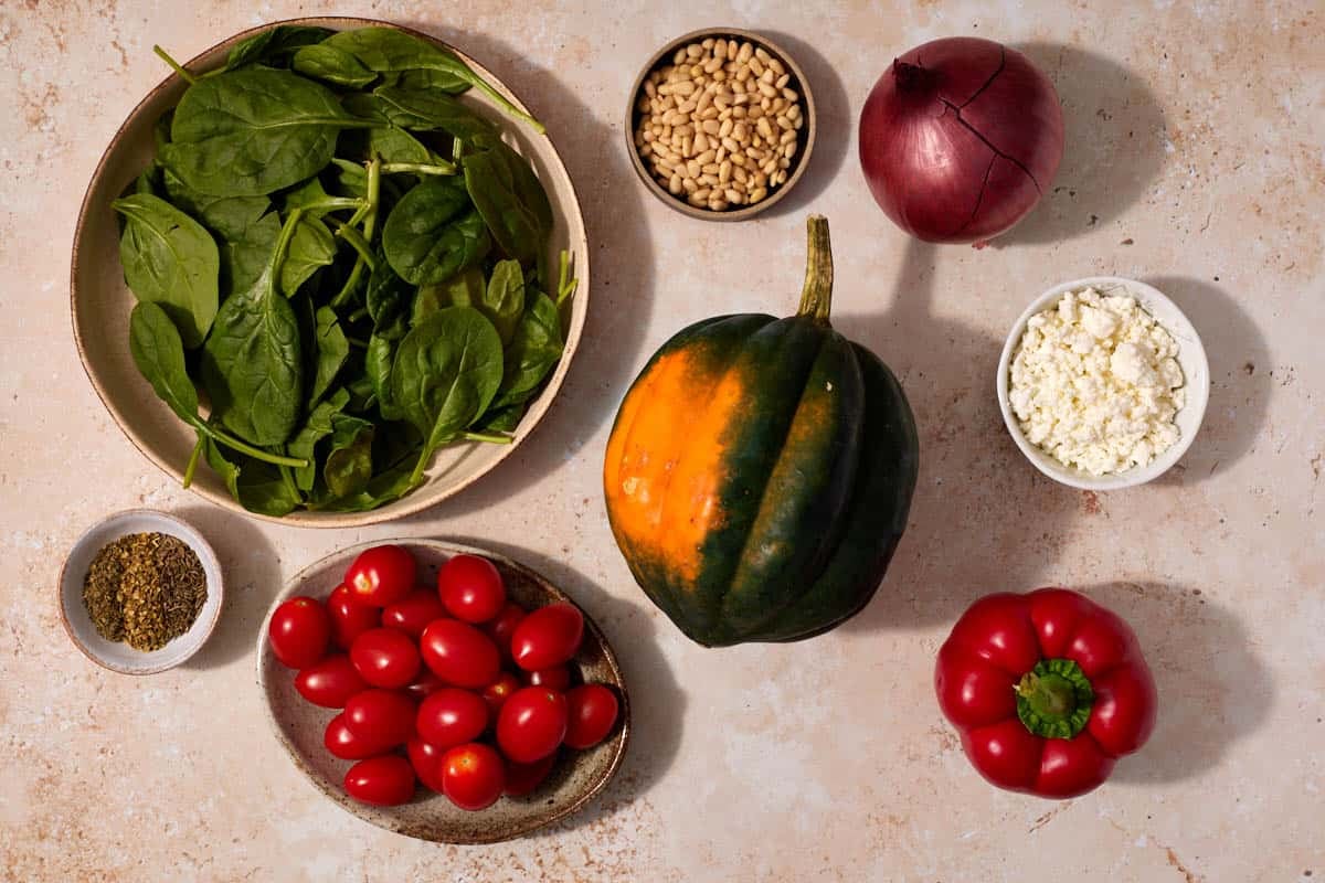 Ingredients for stuffed acorn squash, including pine nuts, acorn squash, orange, bell pepper, red onion, grape tomatoes, extra virgin olive oil, oregano, basil, thyme, salt, black pepper, baby spinach, and goat cheese.