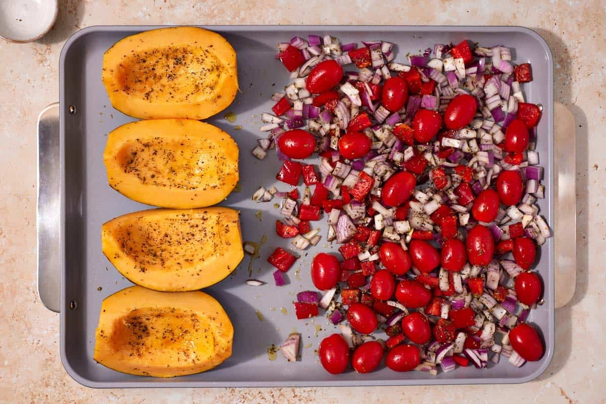 Four acorn squash halves on one side and tomatoes and onions on the other. Both have been seasoned and are drizzled with oil.