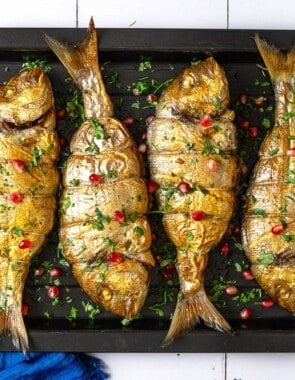 an overhead photo of 4 cooked persian baked fishes garnished with pomegranate seeds and parsley on a sheet pan.