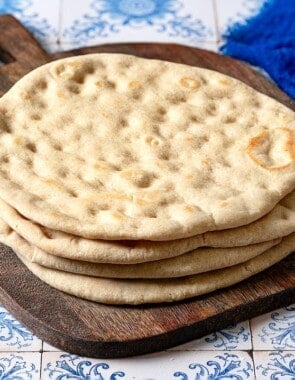 a stack of baked taboon flatbreads on a wooden serving tray next to a small bowl of olive oil and a blue cloth napkin.