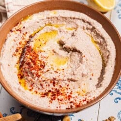 A close up of a tarator sauce in a bowl garnished olive oil and aleppo pepper. Next to this is a kitchen towel, 2 lemon halves, an some walnuts.