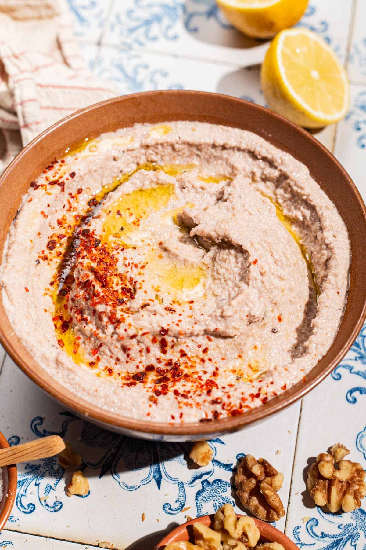 A close up of a tarator sauce in a bowl garnished olive oil and aleppo pepper. Next to this is a kitchen towel, 2 lemon halves, an some walnuts.