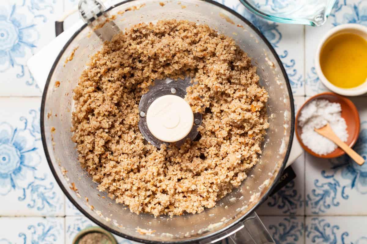 An overhead photo a paste made with bread, garlic, walnuts, and lemon juice in the bowl of a food processor fitted with a blade. Next to this is a measuring cup, and small bowls of olive oil and salt.