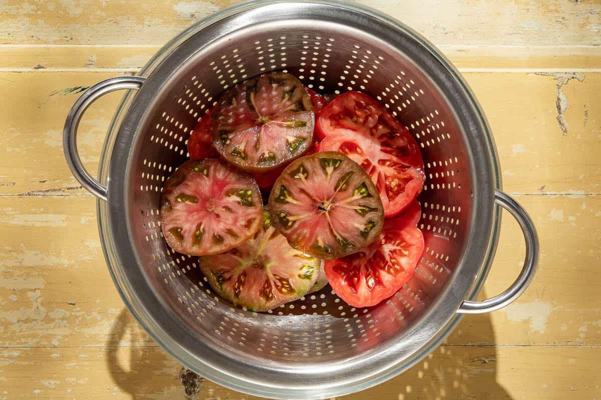 An overhead photo of sliced tomatoes draining in a colander set in a large bowl.