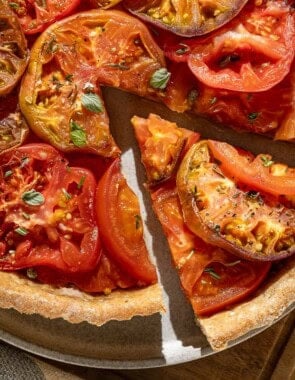 An overhead photo of a tomato tart on a plate on a wooden cutting board with one slice cut from it. Next to this is a kitchen towel and a serving utensil.