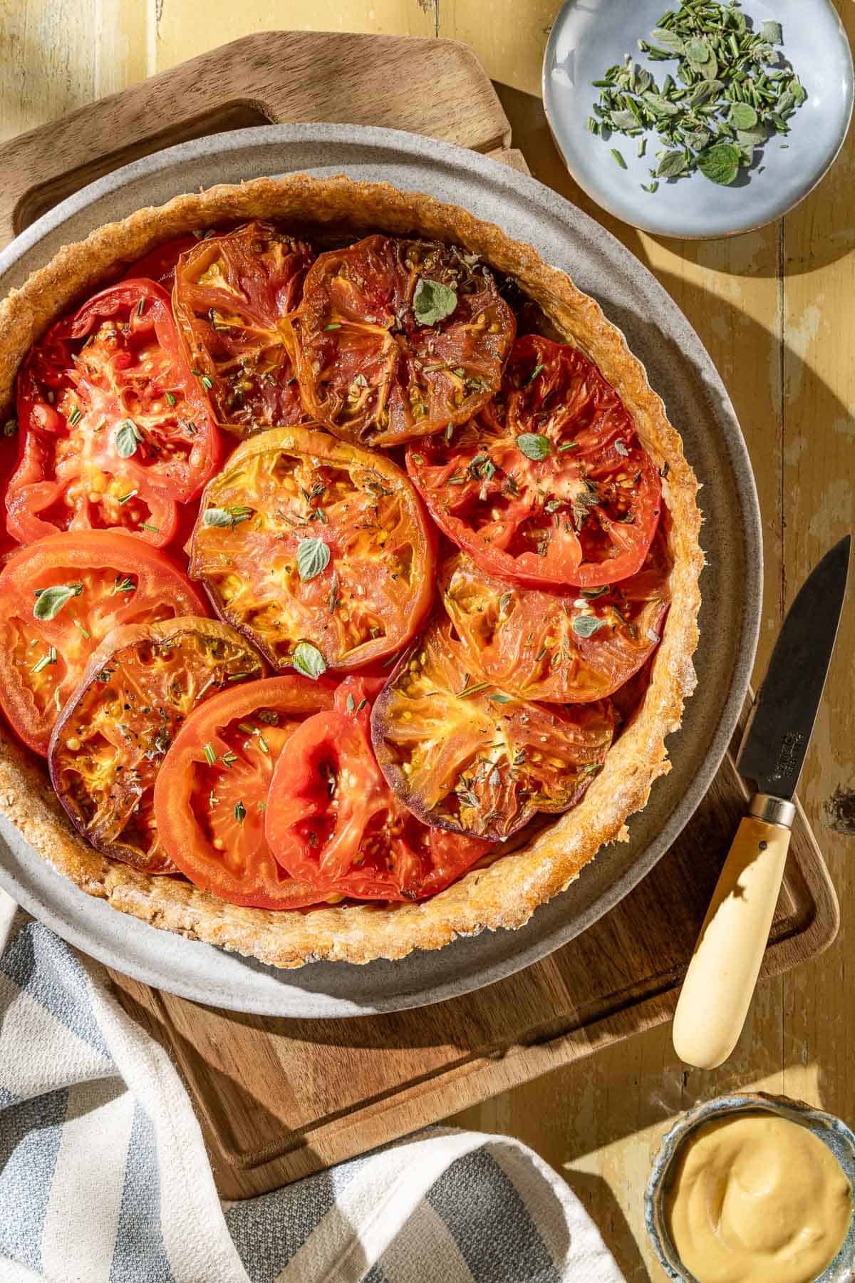 An overhead photo of a tomato tart on a plate on a wooden cutting board. Next to this is a knife, a kitchen towel, a bowl of mustard and a plate of fresh herbs.