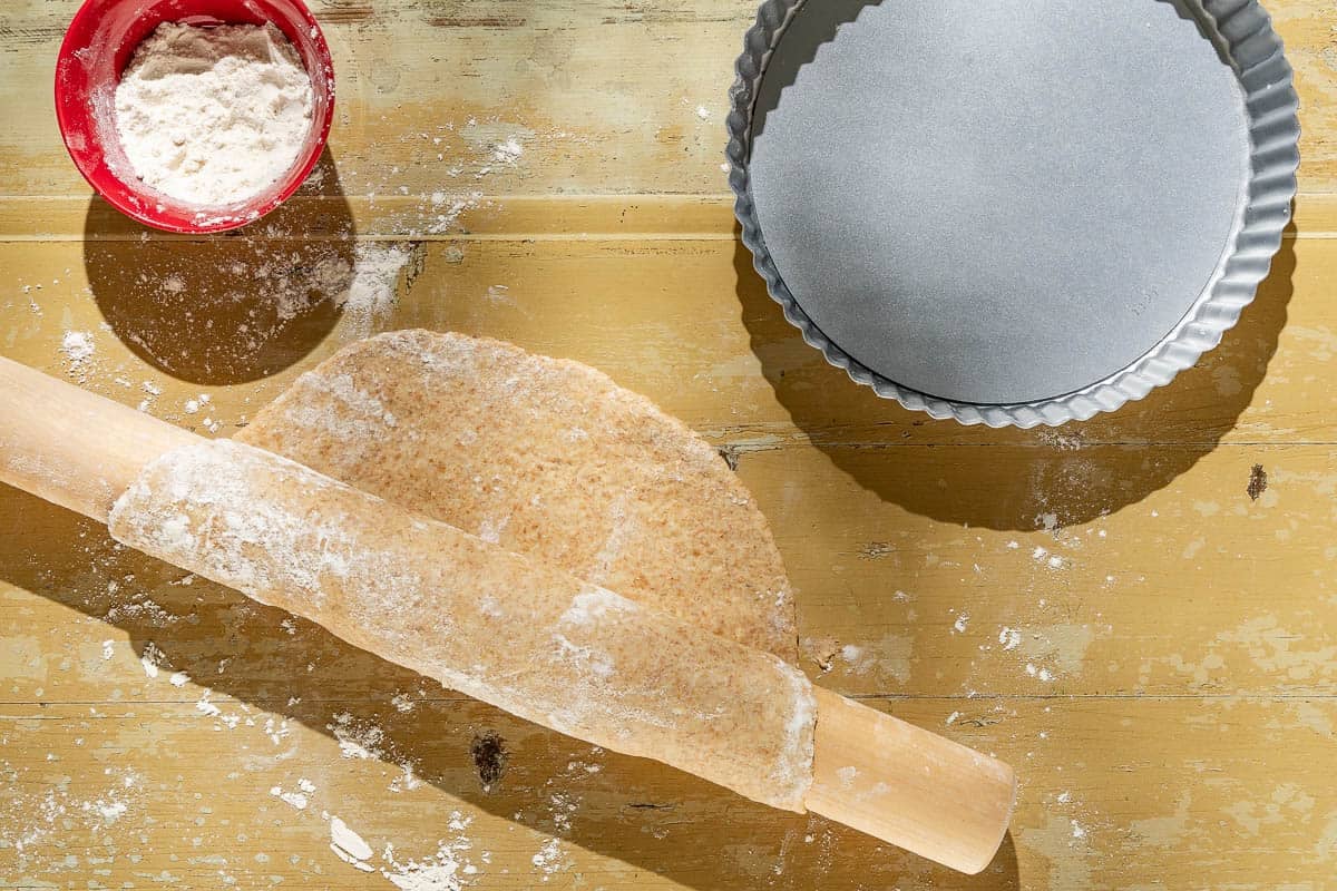 The dough for the tomato tart crust wrapped around a rolling pin. Next to this is a bowl of flour and a tart pan.