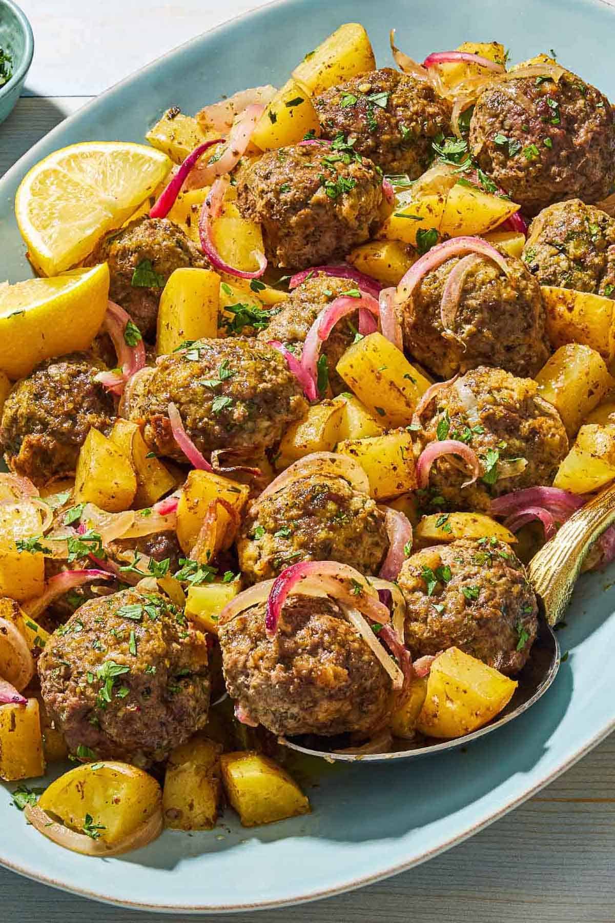 A close up of baked greek meatballs and potatoes on a serving plate with a serving spoon and lemon wedges.