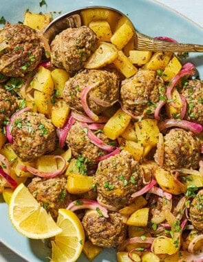 Baked greek meatballs and potatoes on a serving plate with a serving spoon and lemon wedges. Next to this is a small bowl of chopped parsley.