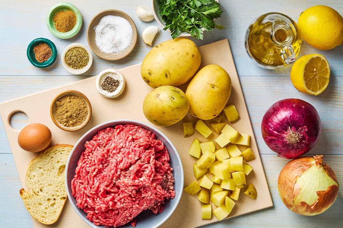 Ingredients for baked greek meatballs and potatoes including ground beef, bread, onions, garlic, egg, parsley, oregano, coriander, cumin, nutmeg, salt, black pepper, gold potatoes red onion, lemon and olive oil.