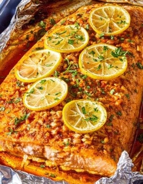 A close up photo of a lemon garlic salmon fillet topped with lemon wheels and parsley on an aluminum foil-lined sheet pan.