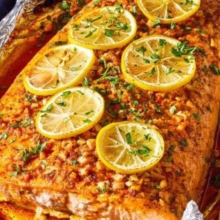 A close up photo of a lemon garlic salmon fillet topped with lemon wheels and parsley on an aluminum foil-lined sheet pan.