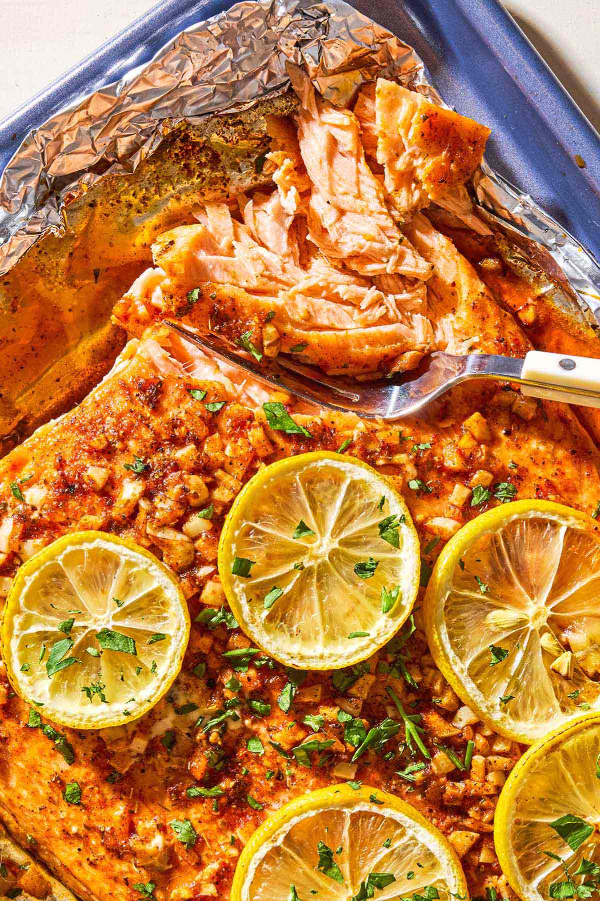 An close up photo of a lemon garlic salmon fillet topped with lemon wheels and parsley on an aluminum foil-lined sheet pan with a fork.