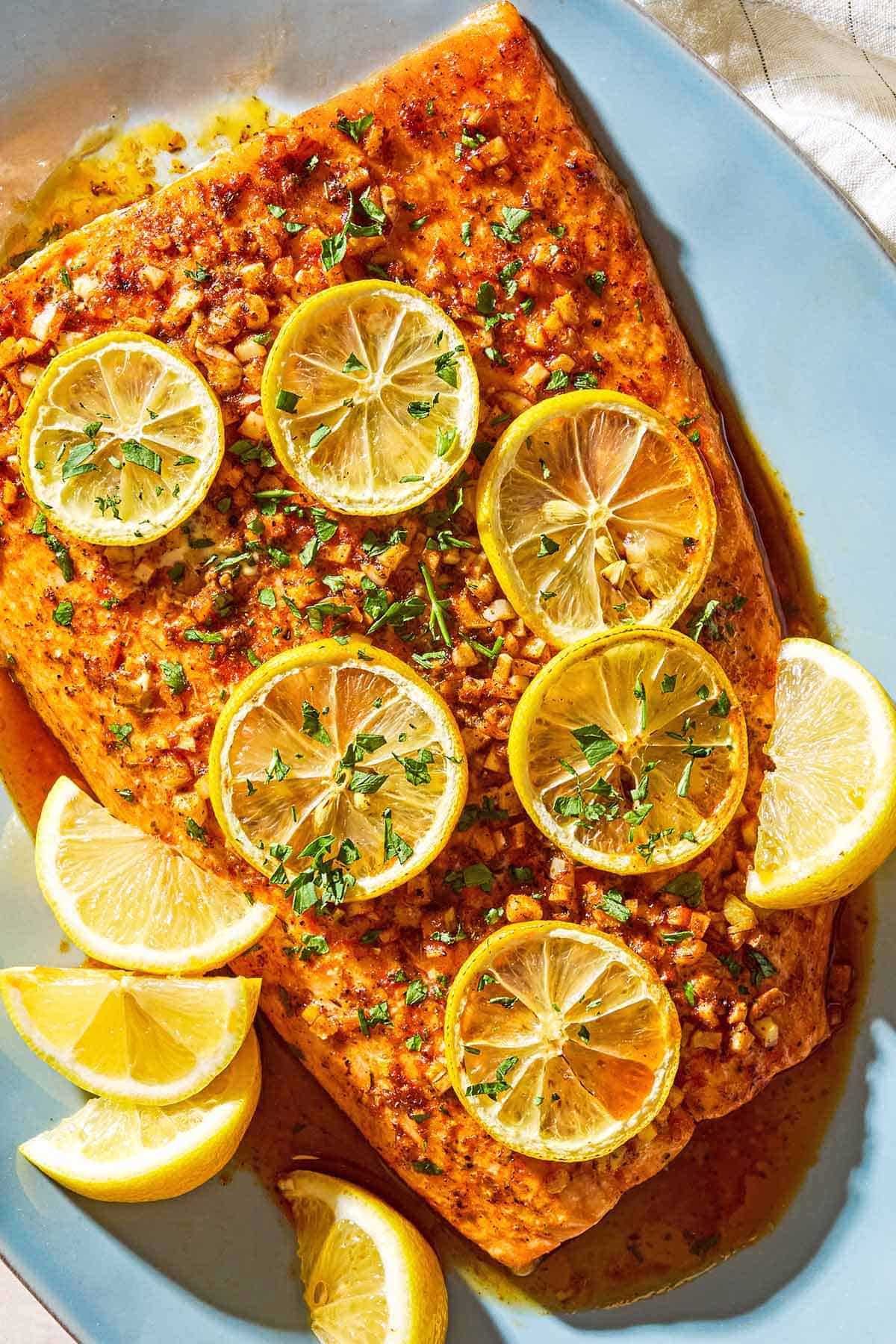 An overhead photo of a lemon garlic salmon fillet topped with lemon wheels and parsley on a serving platter with lemon wedges.