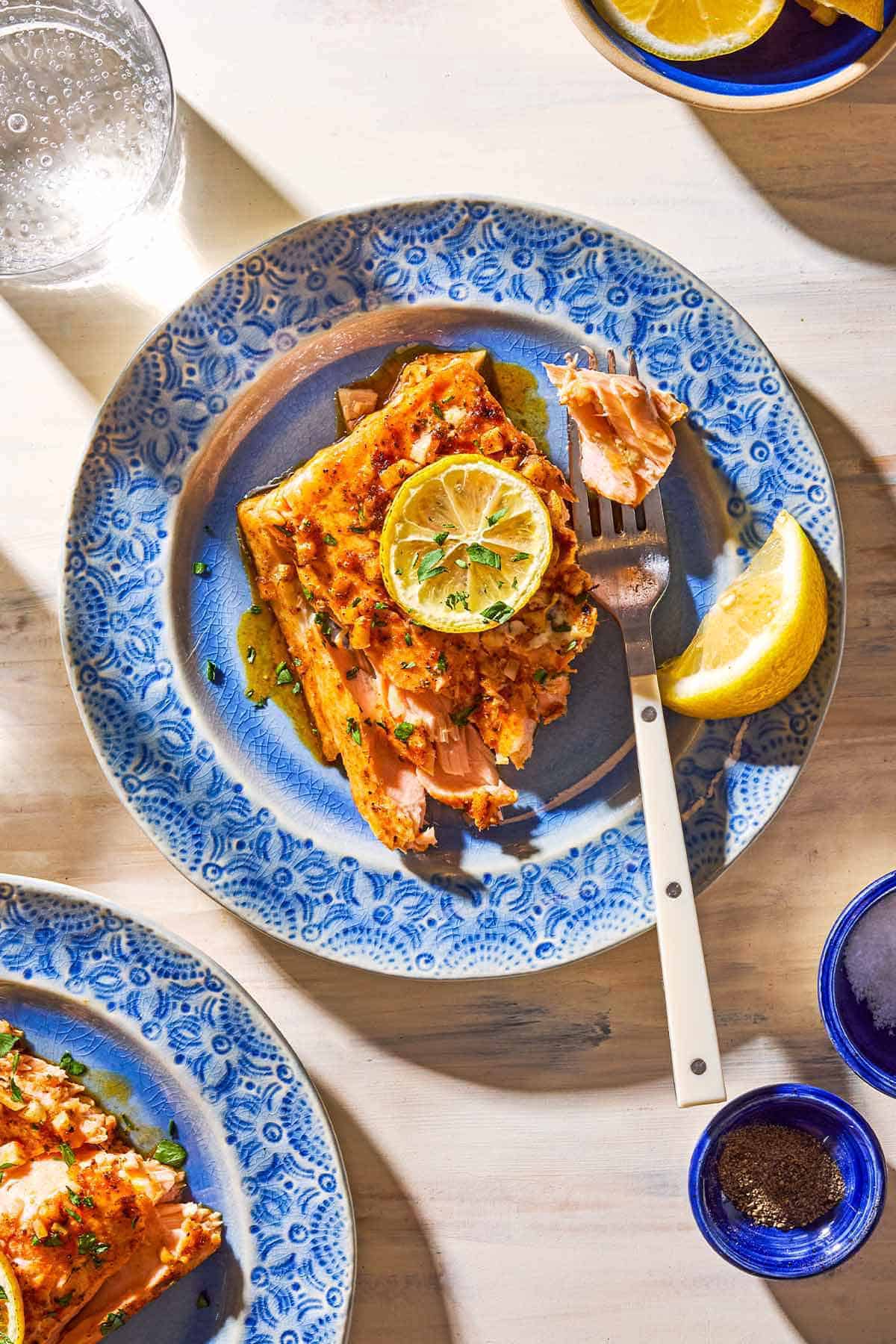 An overhead photo of a serving of the lemon garlic salmon topped with a lemon wheel and parsley on a plate with a fork and a lemon wedge. Next to this is another serving of the salmon on a plate, a glass of water, and small bowls of salt and pepper.