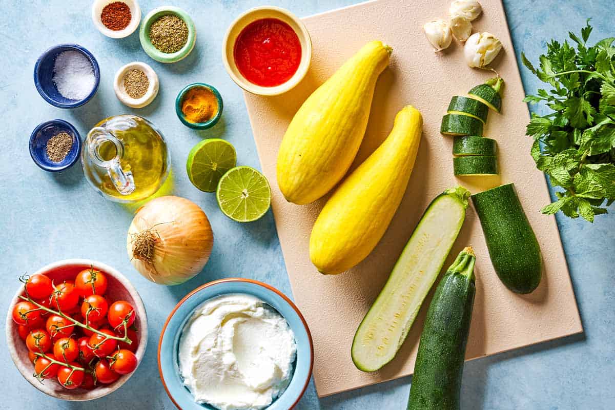 Ingredients for Borani Kadoo including zucchini, olive oil, salt, pepper, onion, garlic, turmeric, oregano, coriander, aleppo pepper, tomato paste cherry tomatoes, lime, fresh herbs, and greek yogurt.