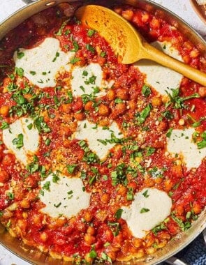 An overhead photo of chickpeas in tomato sauce with a wooden spoon in a skillet. Next this is a plate with crusty bread, a kitchen towel and a small bowl of pecorino romano cheese.