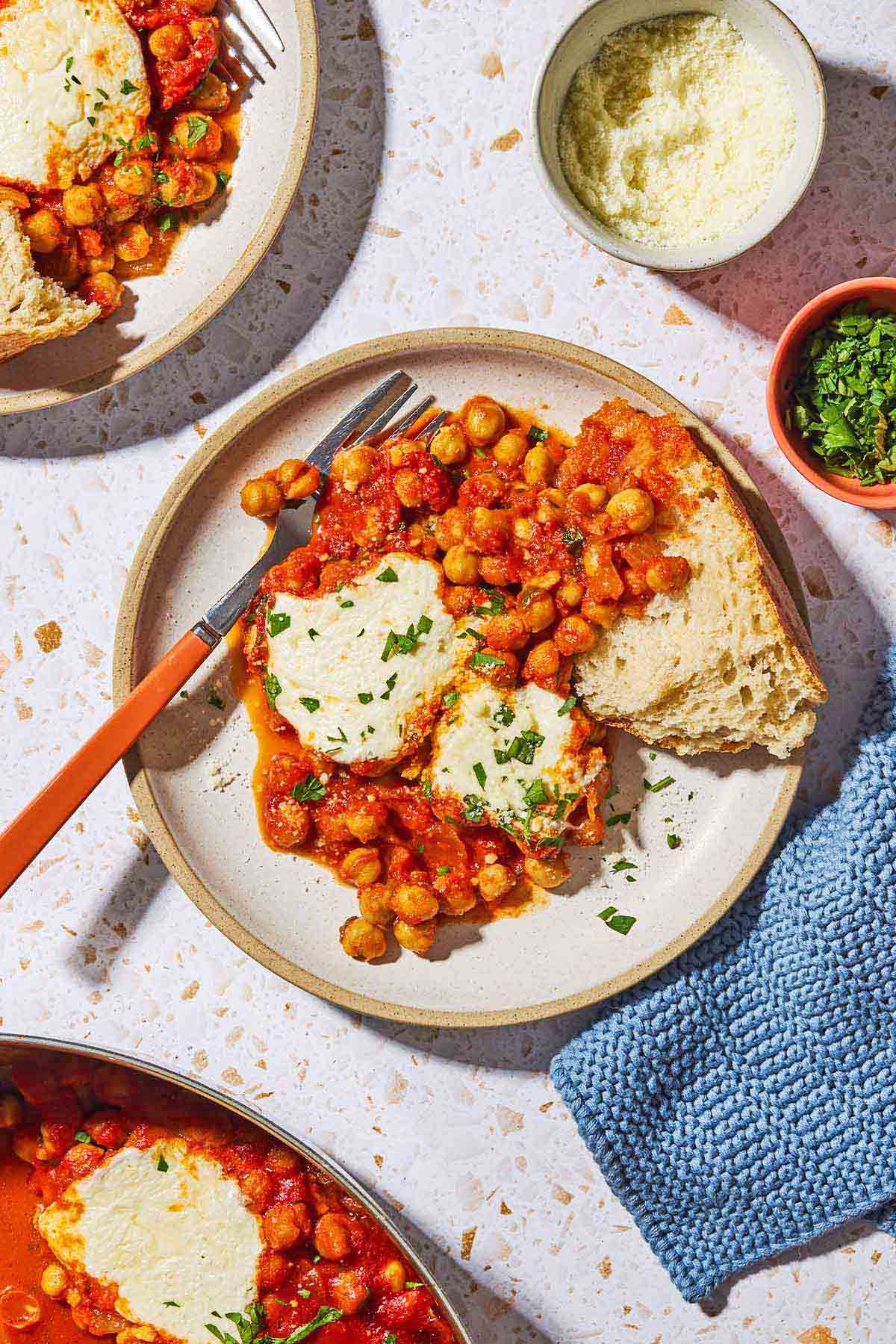 An overhead photo of a serving of chickpeas in tomato sauce on a plate with a fork. Next to this is another serving of chickpeas in tomato sauce on a palate, small bowls of grated pecorino romano cheese and chopped parsley, and a kitchen towel.