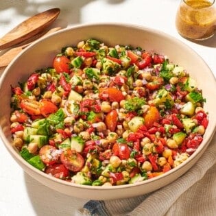 a close up of vegan chickpea salad in a serving bowl next to a wooden serving spoon and fork, a jar of dijon dressing, and a cloth napkin.