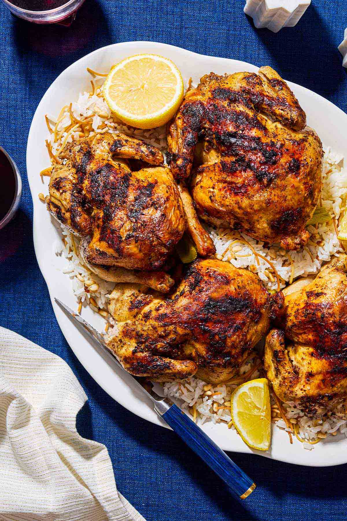 4 roasted cornish hens on a bed of rice on a serving platter with lemon wedges and a knife. Next to this is a cloth napkin.