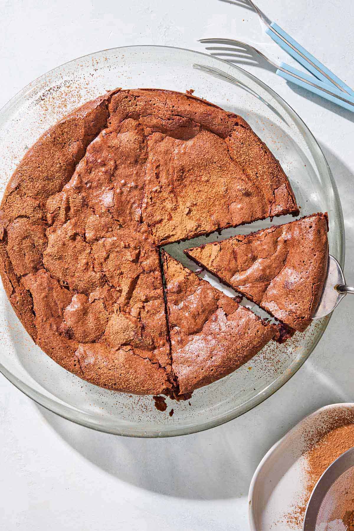 An overhead photo of the flourless chocolate cake with 2 slices cut on a serving platter. Next to this is 2 forks and some cocoa powder in a bowl.