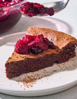 A close up of a slice of flourless chocolate cake topped with berry compote on a plate with a spoonful of the compote being held above it. Behind this is a bowl of the rest of the compote.