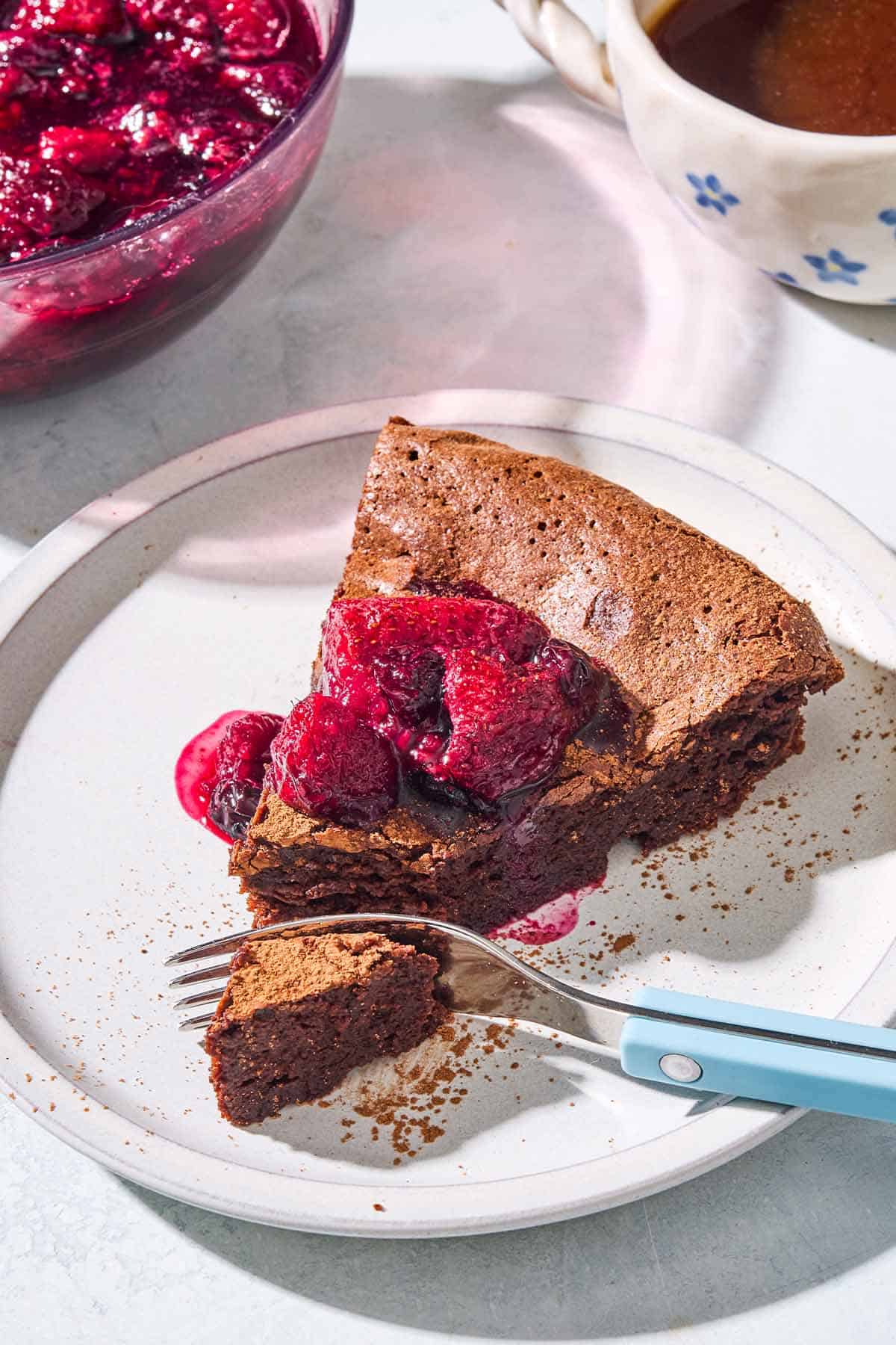 A close up of a slice of flourless chocolate cake topped with berry compote on a plate with a fork. Behind this is a bowl of the compote and a cup of coffee.