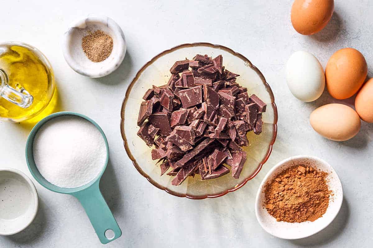 Ingredients for flourless chocolate cake including chopped bittersweet chocolate, olive oil, sugar, ground cardamom, almond extract, eggs, and cocoa powder.