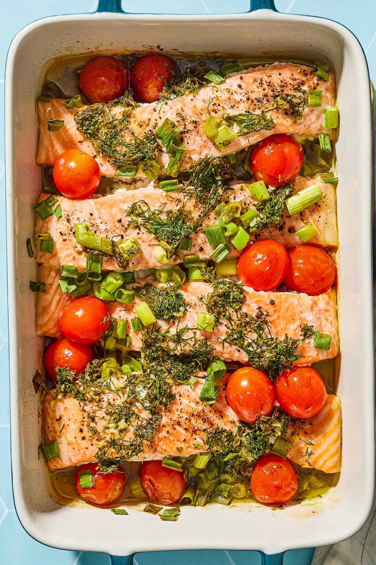 An overhead photo of 4 Greek salmon fillets with cherry tomatoes in a baking dish.