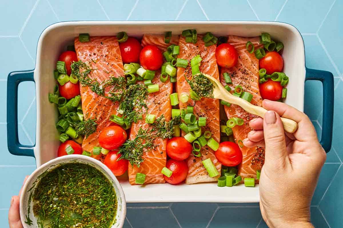 4 uncooked Greek salmon fillets with cherry tomatoes in a baking dish being topped with a Greek-style sauce.