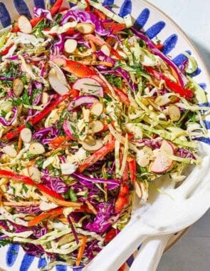 An overhead photo of cabbage salad in a serving bowl with serving utensils. Next to this is a small container of the dressing and a stack of 2 plates.