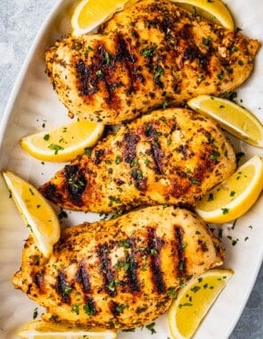 Overhead shot of three grilled chicken breasts that have been marinated in Greek chicken marinade and are on a platter with lemon wedges and a sprinkle of parsley.