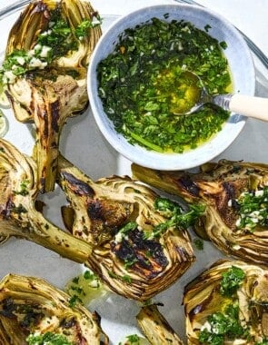 Grilled artichoke quarters drizzled with marinade on a serving platter along with some of the marinade in a bowl with a spoon.