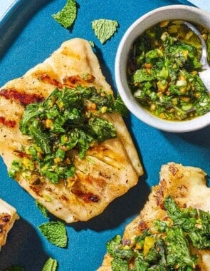 An overhead photo of 2 grilled cod filets topped with pistachio-herb salsa on a serving platter along with a small bowl of the salsa and a spoon.