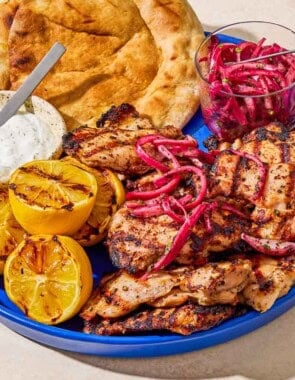 Grilled sumac chicken thighs on a serving platter with pickled red onions, grilled lemon halves, tzatziki sauce, and pita bread.