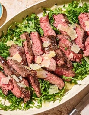 An overhead photo of beef tagliata topped with shaved parmesan on a bed of arugula on a serving platter. Next to this is a small bowl of salt and a serving fork.