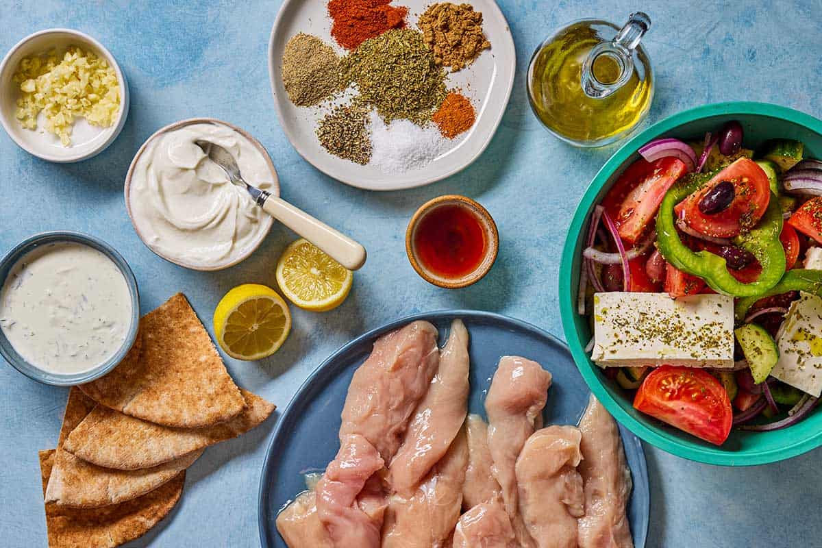 Ingredients for gyro bowls including chicken tenderloins, greek yogurt, lemon, red wine vinegar, garlic, oregano, paprika, cumin, coriander, cayenne pepper, salt, pepper, tzatziki sauce, a greek salad and pita bread.