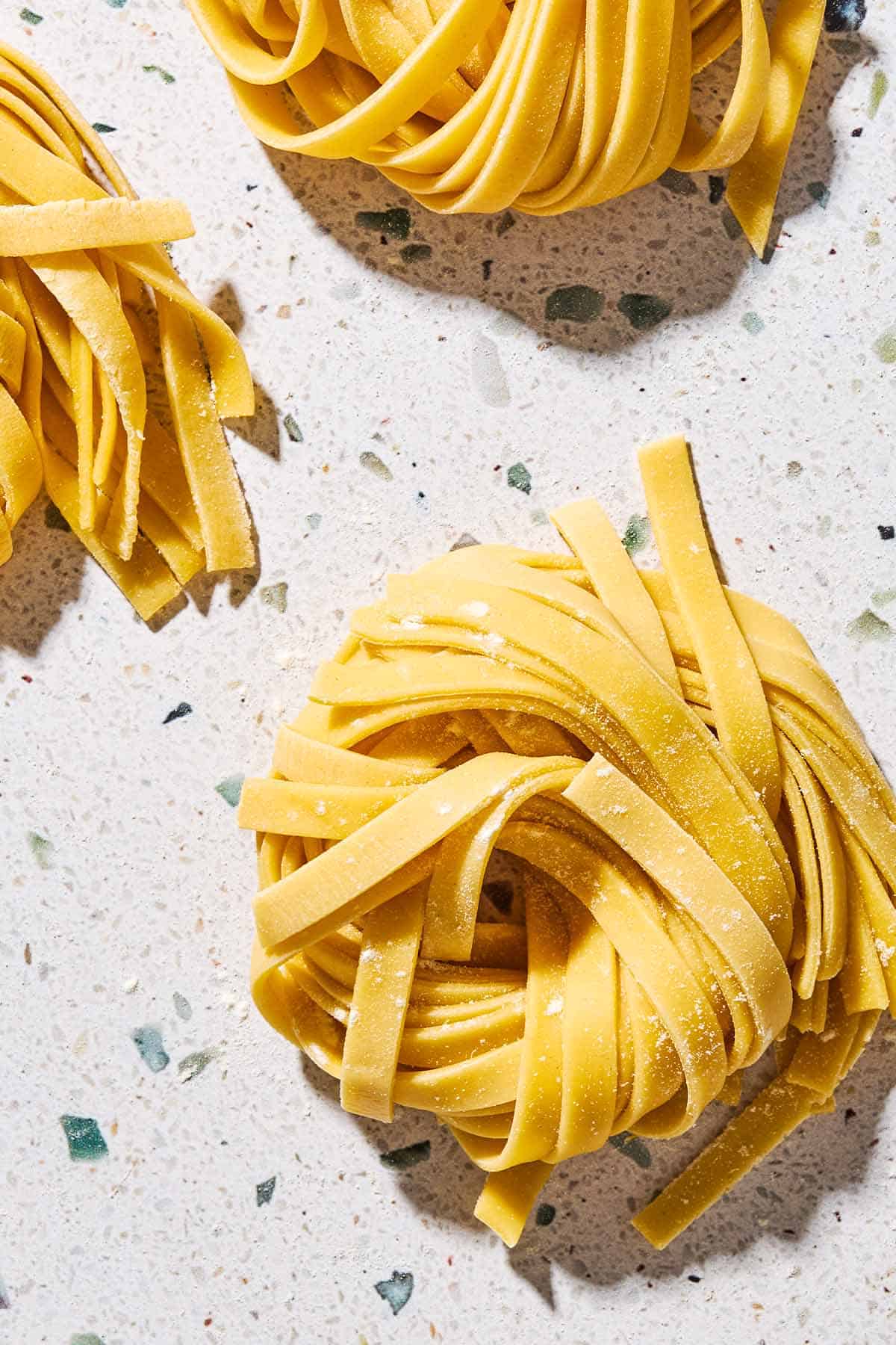 An overhead close up photo uncooked homemade pasta noodles formed into 3 nest shapes.