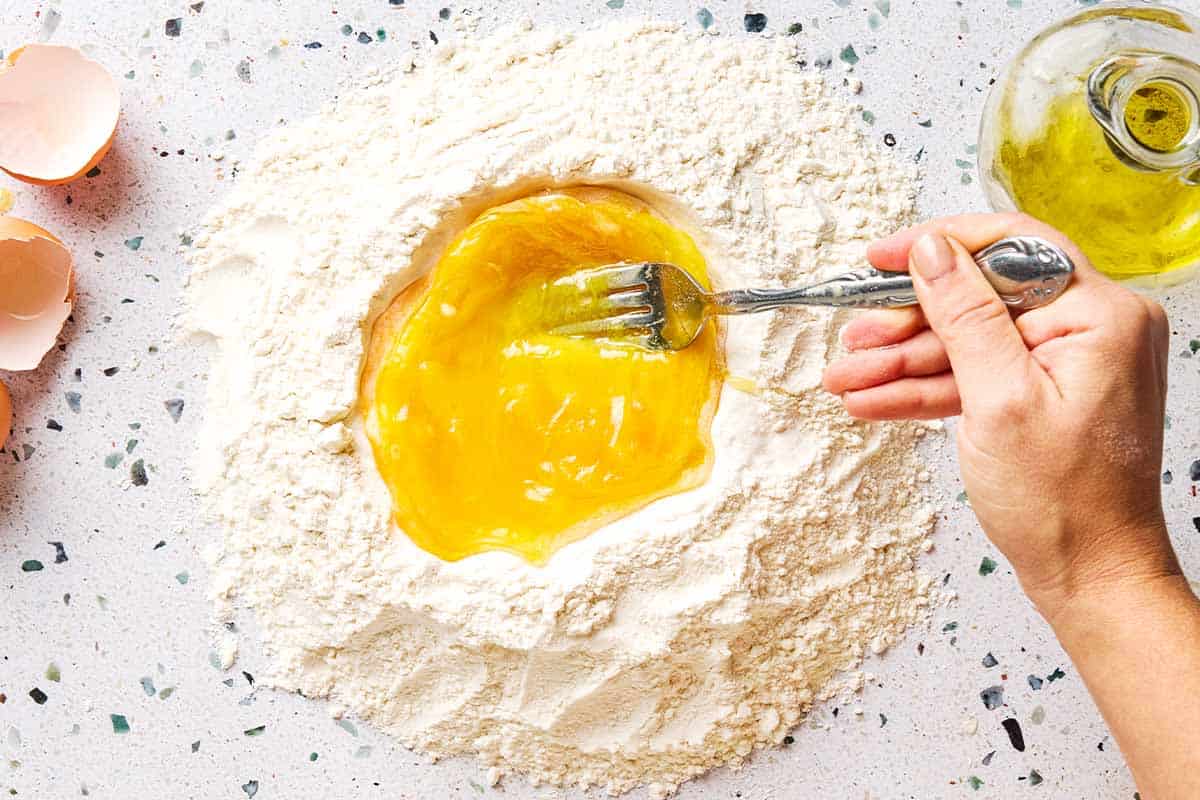 Eggs and olive oil being mixed together in the well of a mound of flour. Next to this is a small jug of olive oil and some egg shells.