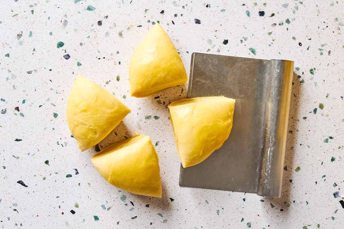 An overhead photo of 4 equal portions of a homemade pasta dough ball on a table with a stainless steel scraper.