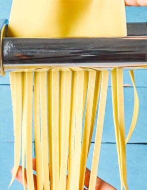 A close up noodles being cut from sheet of homemade pasta dough that's being run through a pasta machine.