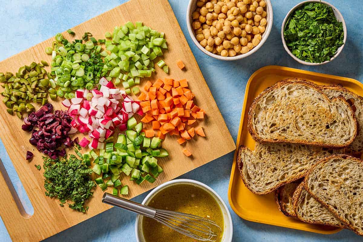 ingredients for vegan chickpea salad sandwich including chickpeas, slices of bread, celery, carrot, persian cucumber, radishes, green onions, kalamata olives, green olives, parsley, mint, and dijon vinaigrette.