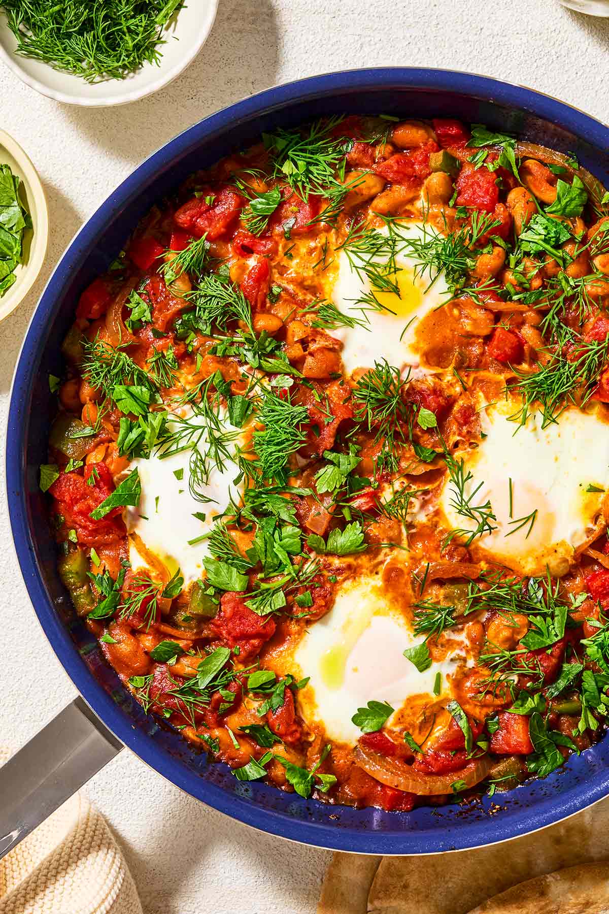 An overhead photo of white bean shakshuka garnished with dill and parsley in a skillet. Next to this are small bowls of dill and parsley.