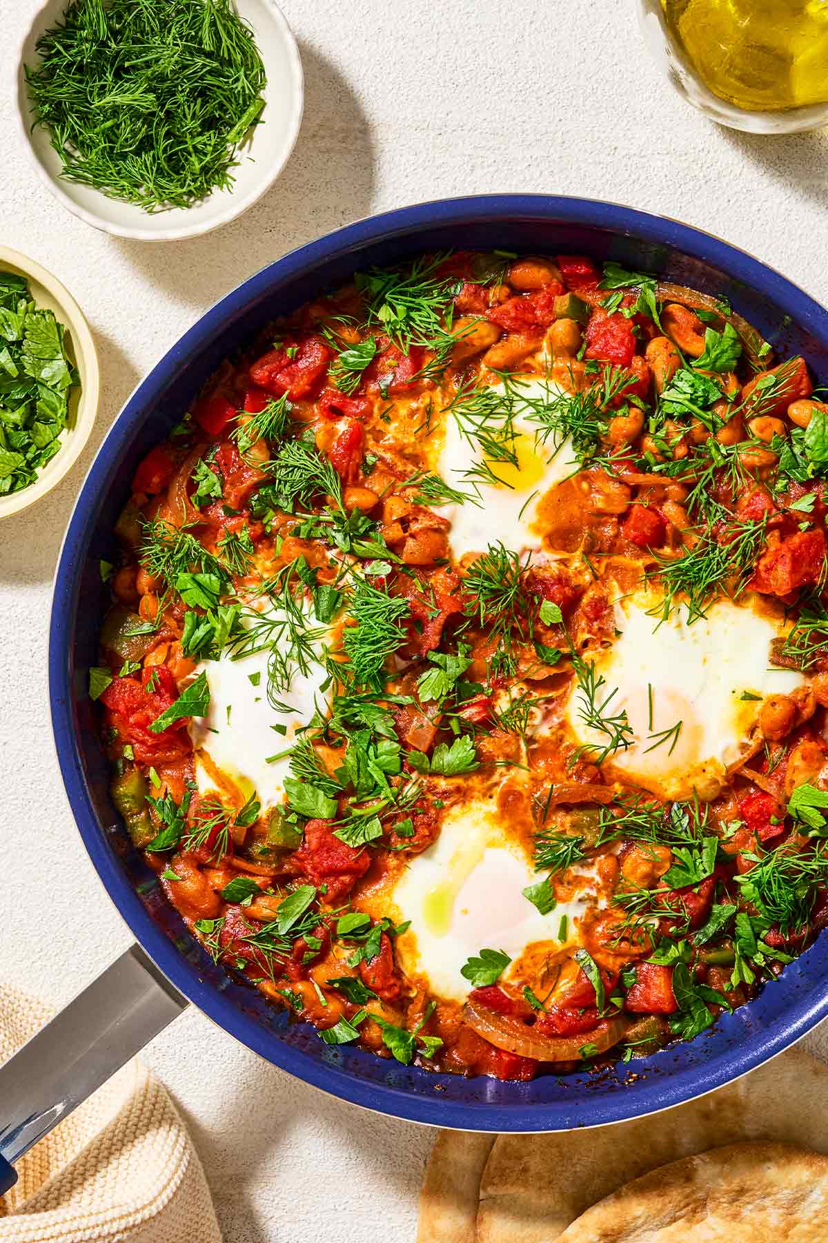 An overhead photo of white bean shakshuka garnished with dill and parsley in a skillet. Next to this are small bowls of dill and parsley, a cup of olive oil, and a stack of pita bread.