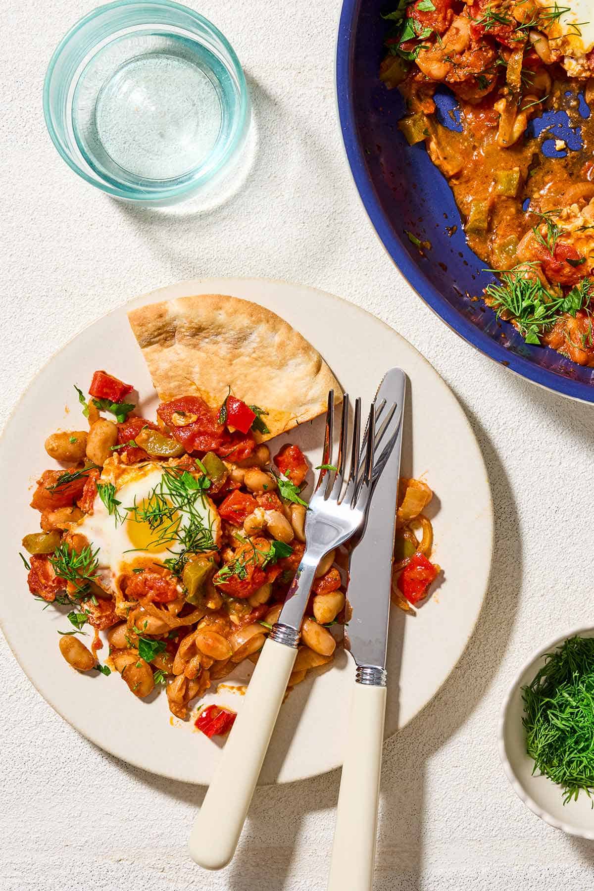 An overhead photo of a plate with a serving of the shakshuka and a knife and fork. Next to this is the rest of the shakshuka in a skillet, a glass of water and a small bowl of dill.