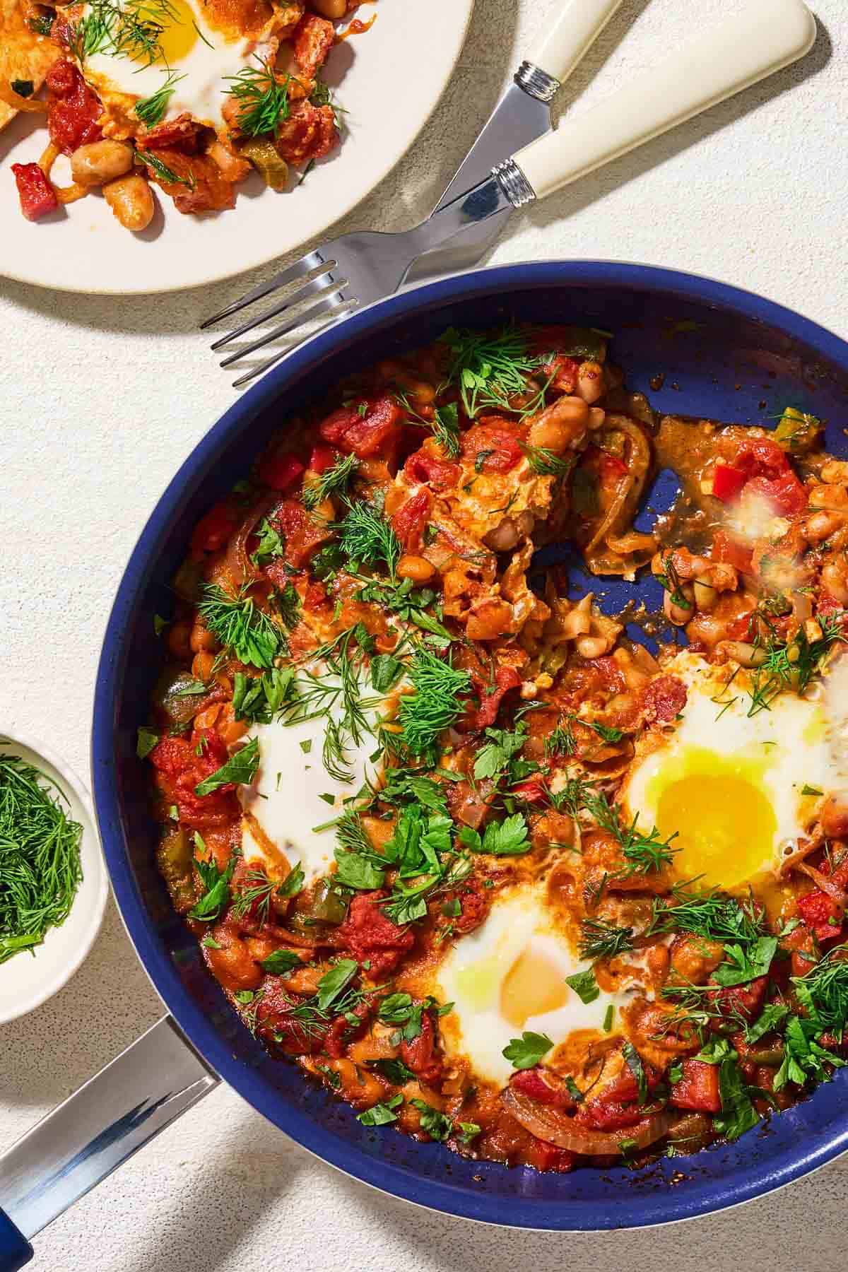 white bean shakshuka garnished with dill and parsley in a skillet. Next to this is a plate with a serving of the shakshuka, a knife and fork, and a small bowl of dill.