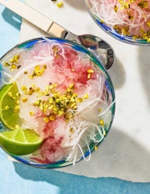 An overhead photo of the faloodeh in a bowl topped with the cherry syrup, chopped pistachios and lime wedges. Next to this is a spoon, 2 lime wedges, and another bowl of the faloodeh.