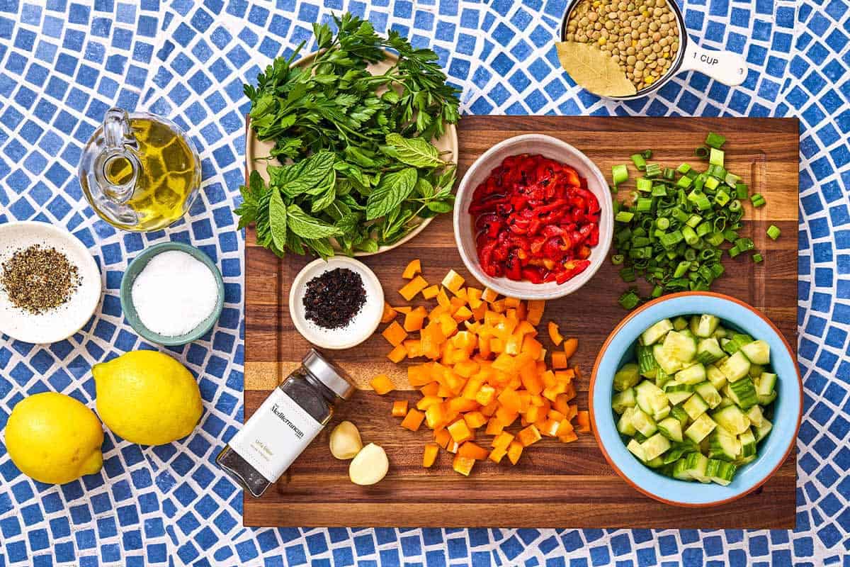 Ingredients for lentil salad including lentils, cucumber, a bay leaf, bell peppers, green onions, parsley, mint, lemons, garlic, salt, pepper, urfa biber and olive oil.