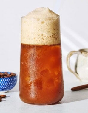 A close up of Freddo Espresso in a glass next to a bowl of coffee beans, a small pitcher and a spoon.