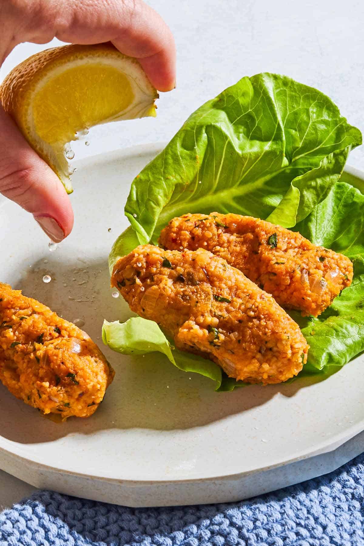 A shot of Mercimek Köftesi, Turkish lentil "meatballs" on a plate with lettuce and a hand above it squeezing lemon over it. 